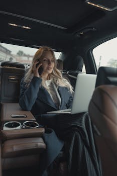 Confident businesswoman making a call and working on a laptop in a luxury car interior.